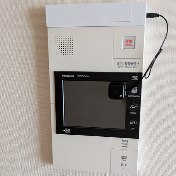 Panasonic製（型番VGD18342W）のオートロック対応インターホンの液晶部分に、ワイヤレスチャイム用の光センサーが貼り付けられた状態。 （液晶画面の右側に、黒い小さな光センサーが取り付けられ、上部から細いケーブルが伸びている写真。）
