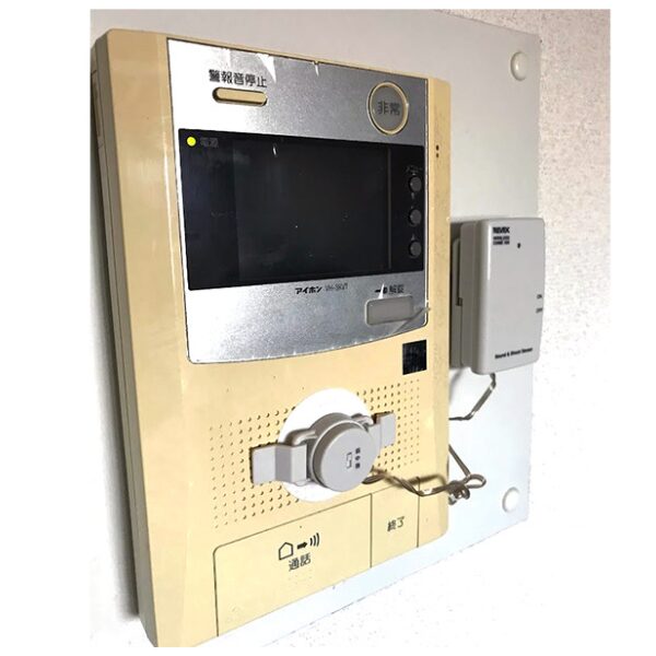 モニター付きインターホン親機に音・振動センサーが取り付けられた状態の設置例写真。センサー部が親機のスピーカー部分に、センサー本体が右横の壁面に設置されている。