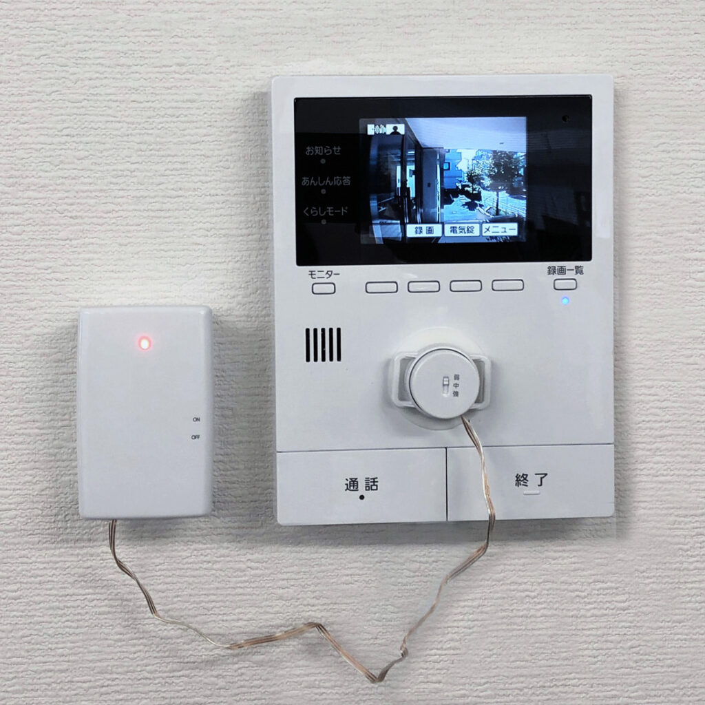 壁に取り付けられたドアホン(テレビドアホン)のモニター親機と、それに接続された外部センサーの画像。モニター画面には玄関先のような映像が表示されており、「お知らせ」「あんしん応答」「くらしモード」といったメニューや、「モニター」「録画一覧」「通話」「終了」といった操作ボタンが見える。モニター下部には音・振動センサーが接続され、左隣には赤いランプが点灯した電池ケースが置かれている。